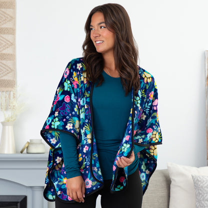 Woman in a cozy setting wearing a blue fleece, pocketed shawl, patterned with paws, flowers and foliage. The edges are lined with black piping, wrapped around her shoulders.