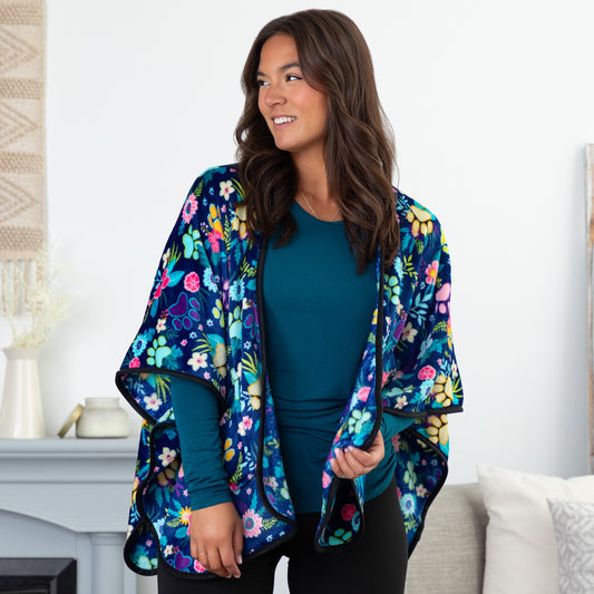 Woman in a cozy setting wearing a blue fleece, pocketed shawl, patterned with paws, flowers and foliage. The edges are lined with black piping, wrapped around her shoulders.