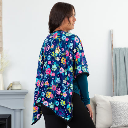 Woman in a cozy setting wearing a blue fleece, pocketed shawl, patterned with paws, flowers and foliage. The edges are lined with black piping, wrapped around her shoulders. She is turned away showing the continuation of the floral pattern on the back of the wrap.