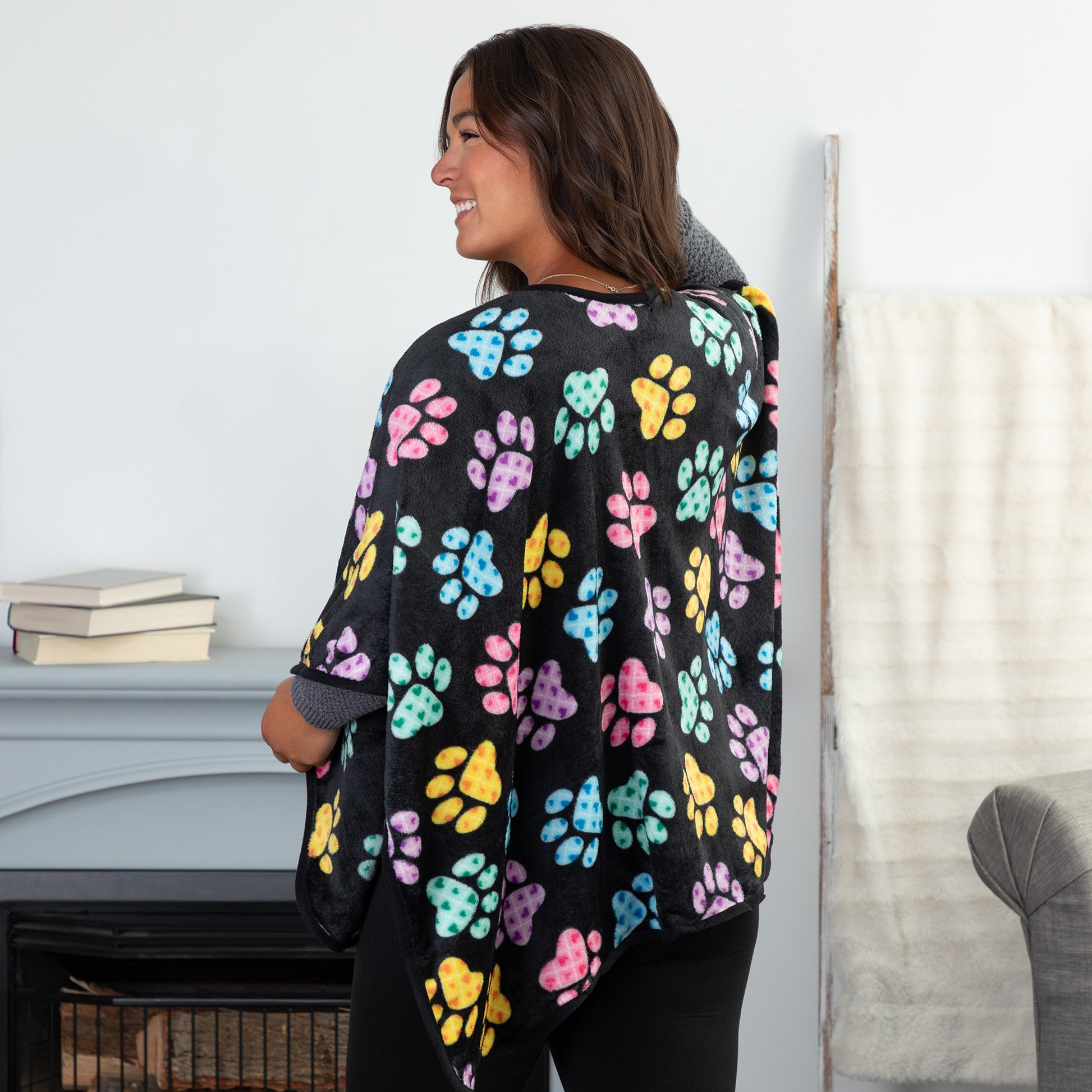 A woman standing in a cozy living room. She is wearing a black fleece, pocketed shawl, patterned with pink, blue, yellow, green, and purple argyle paw prints. She is facing away to show the continuation of pattern on the back of the wearable blanket wrap.