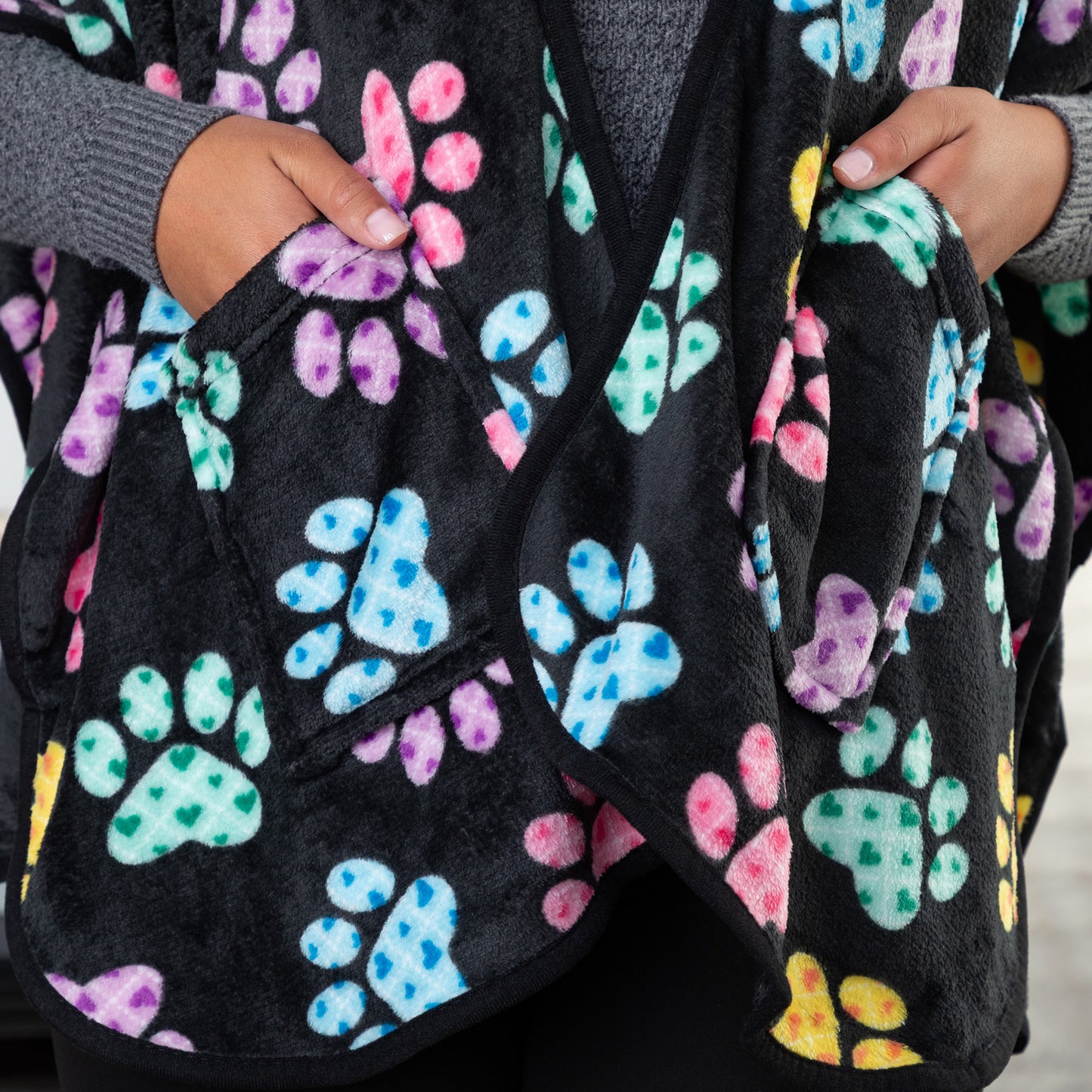 Hands in the two pockets of a black fleece wearable blanket shawl patterned with pink, blue, yellow, green, and purple argyle paw prints.