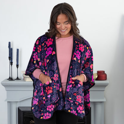 A woman with her hands in her pockets standing in a cozy living room. She is wearing a deep blue based fleece, pocketed shawl, patterned with pink, red and purple paw prints paired with yellow, red, pink, and purple hearts. 