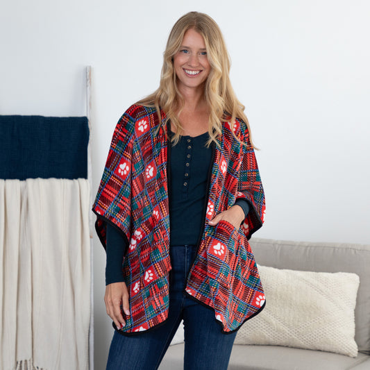 A smiling blonde woman stands indoors wearing a red and navy plaid poncho-style cardigan with white paw prints over a dark top and jeans. A light gray sofa and cream pillow are behind her, with a draped textile on a stand to the left.