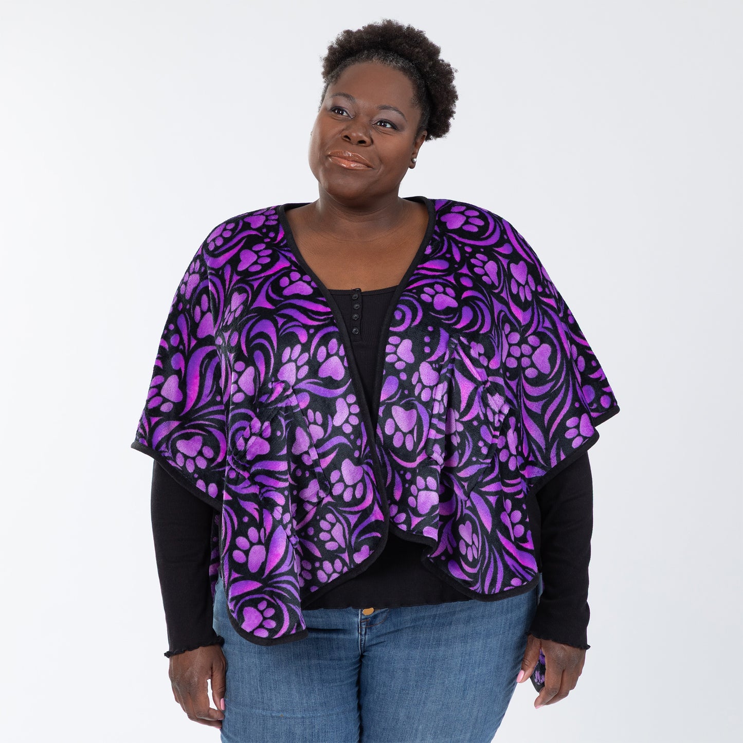 Woman wearing a fleece, pocketed shawl, patterned with purple paw prints and swirls and black piping, wrapped around her shoulders. Available in extended and plus size.