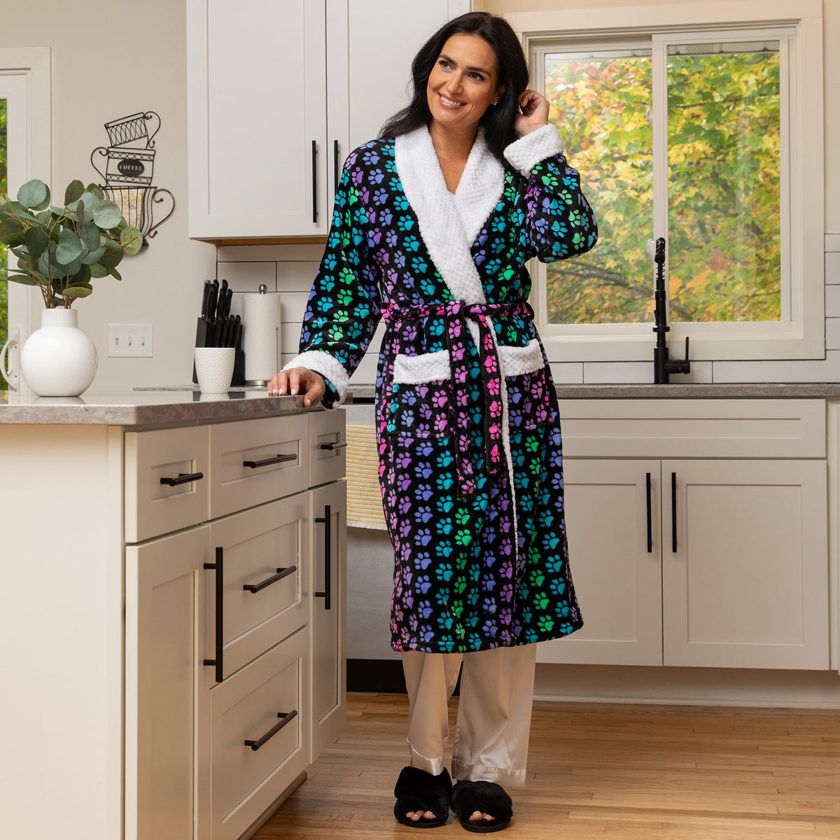 A woman wearing a colorful paw print robe with white trim stands in a bright kitchen, holding a phone to take a mirror selfie. The robe features a vibrant pattern of purple, green, and blue flowers on a dark background, with plush white collar, cuffs, and pocket trims.