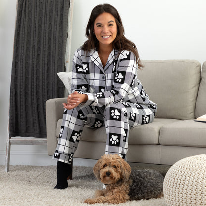 A woman sitting next to her pet dog in a cozy living room setting. She is wearing a matching pajama set of white and black, plaid, button-up, long-sleeve top patterned with paw prints and pants of the same color and pattern. 