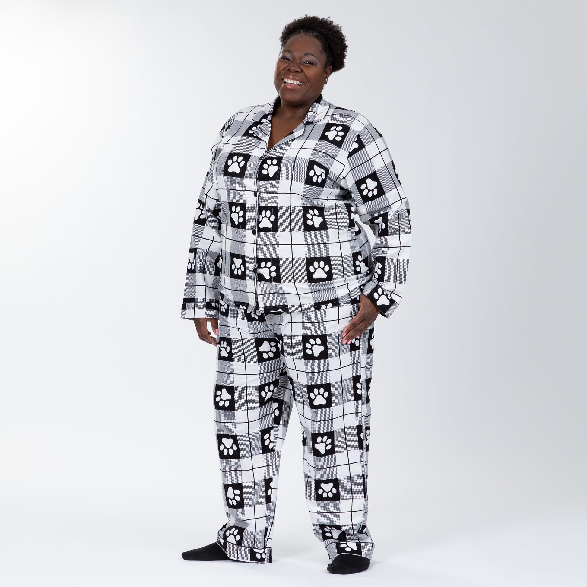 A woman wearing a matching pajama set of white and black, plaid, button-up, long-sleeve top patterned with paw prints and pants of the same color and pattern. Hand in pocket of pajamas. Plus and extended size available. 
