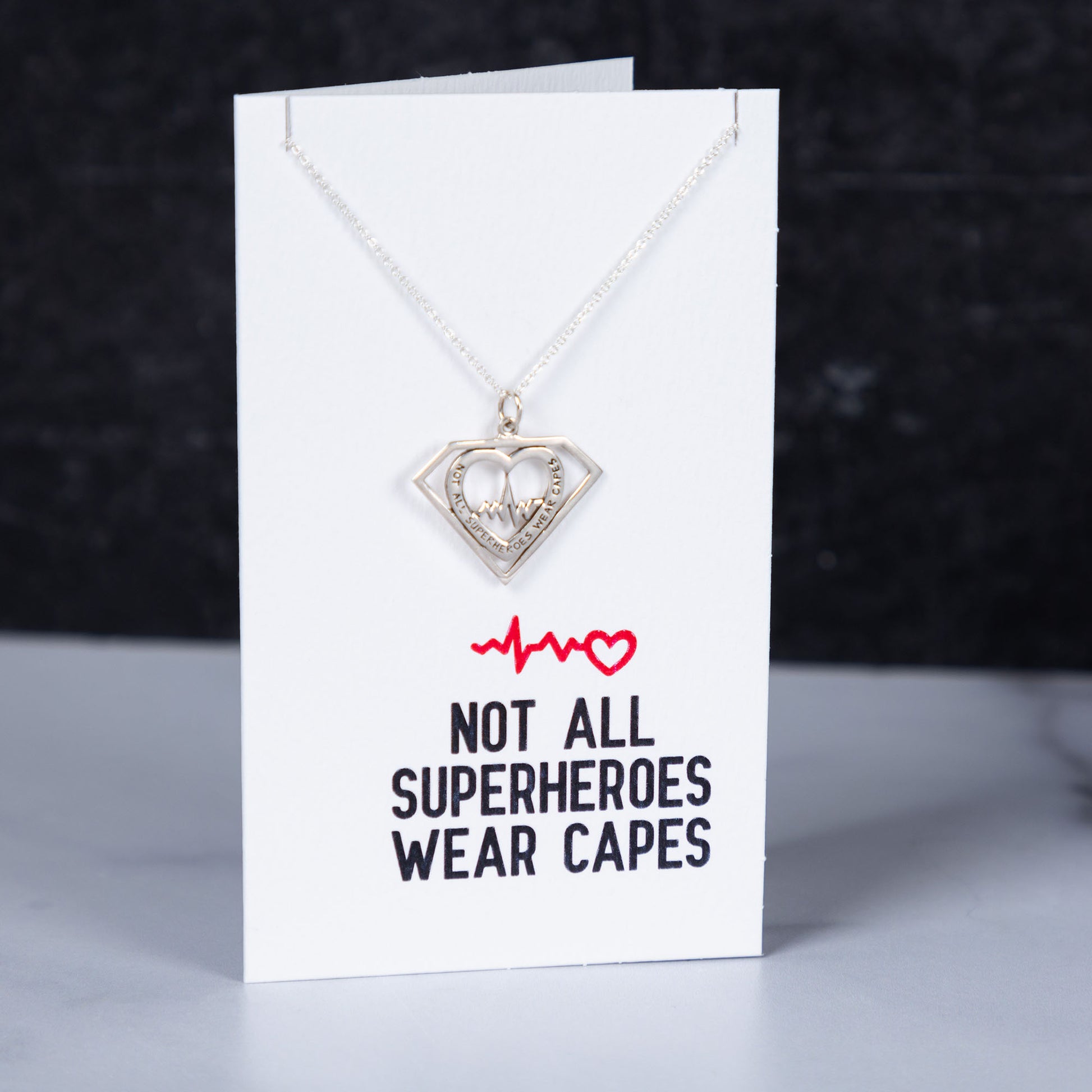 Necklace with heart pendant on a card with text 'Not all superheroes wear capes' against a dark background
