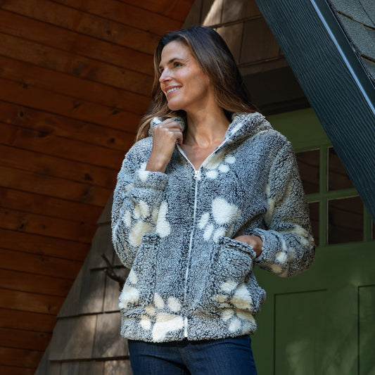 Woman wearing a gray fleece jacket with paw prints standing in front of a wooden cabin.