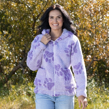 A woman wearing a light purple fuzzy zip-up hoodie with darker purple paw print patterns, standing outdoors in front of a blurred background of green and orange foliage.