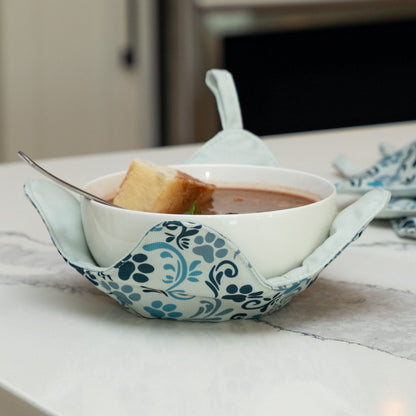 A set of four bowl cozies with various patterns, including a paw print and floral design, paw serenade, with a bowl cozy holding a warm bowl of soup and bread in a comfy kitchen setting