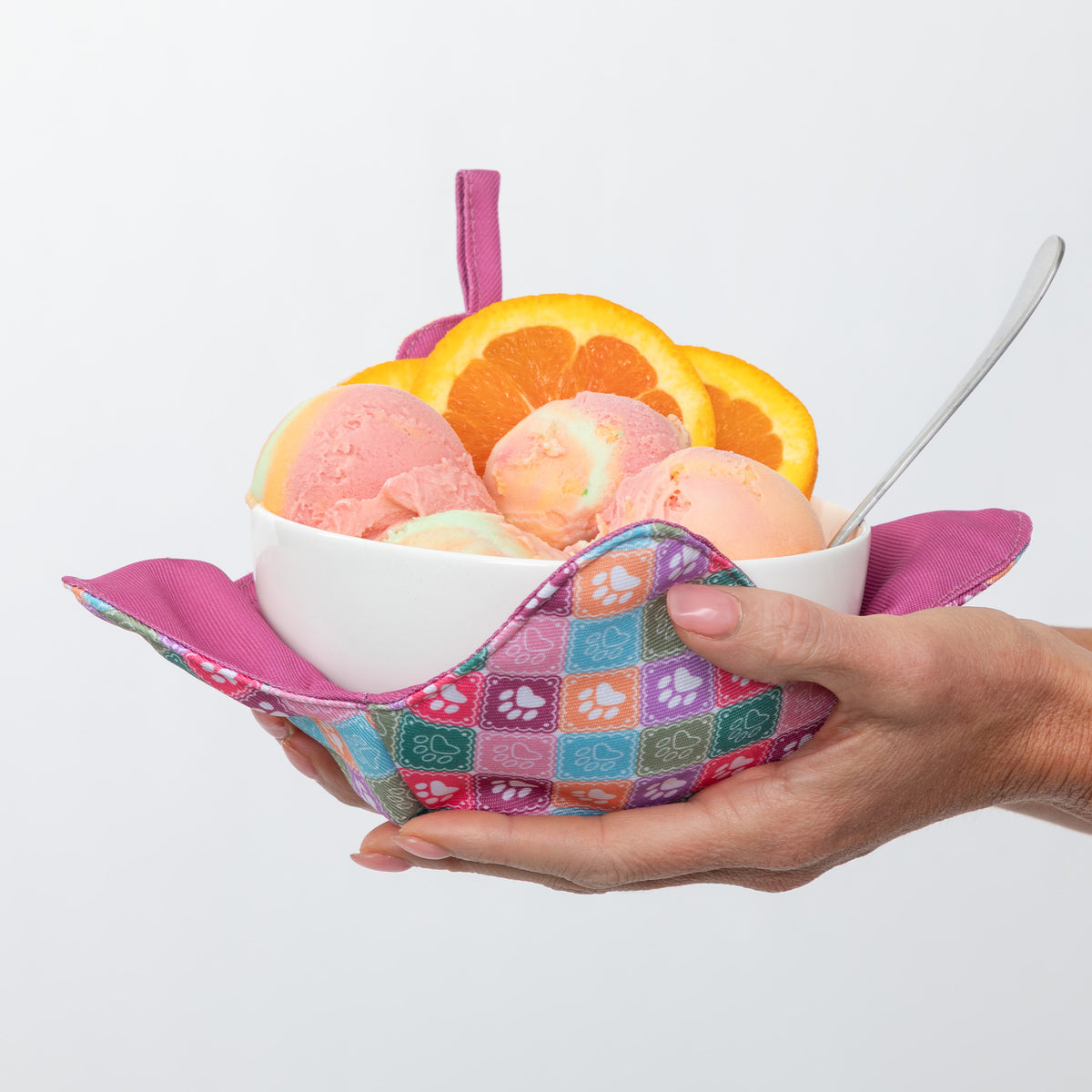 A white bowl filled with pink and peach ice cream or gelato, topped with orange slices, held in a person's hands inside a colorful patterned fabric scoop or holder with a pink handle, against a light gray background.