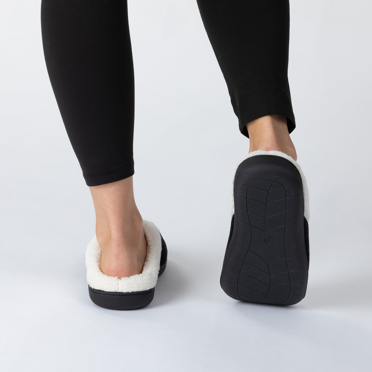 A person wearing black leggings and black slip-on slippers with white sherpa lining, shown from the back against a light gray studio background.