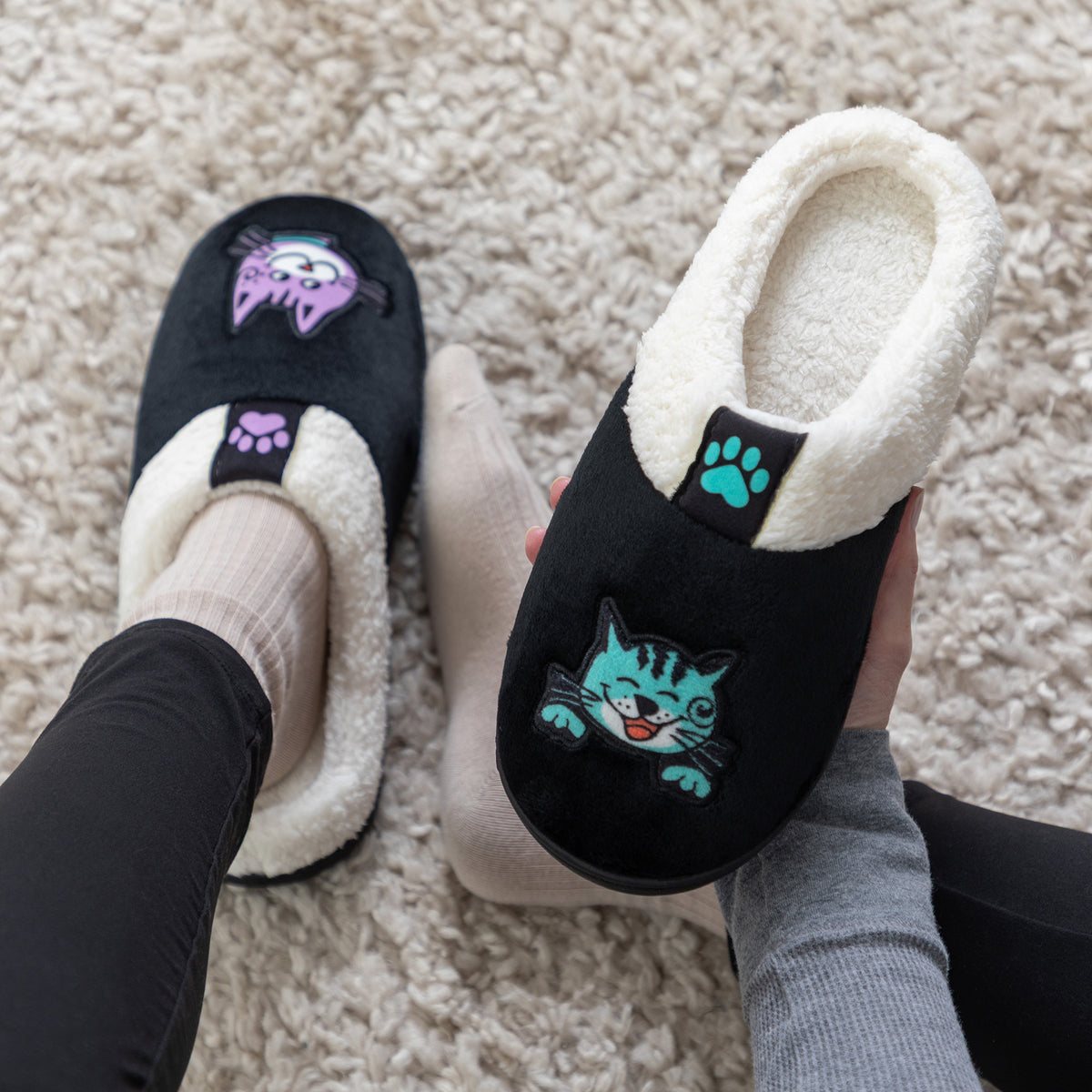 Black and white sherpa slippers with purple and teal cat on top on models feets with a soft beige rug in the background