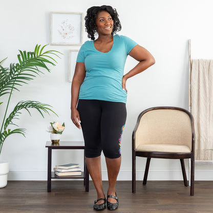 Woman in a teal shirt and black legging shorts to the knee, standing in a room with a chair and plant.