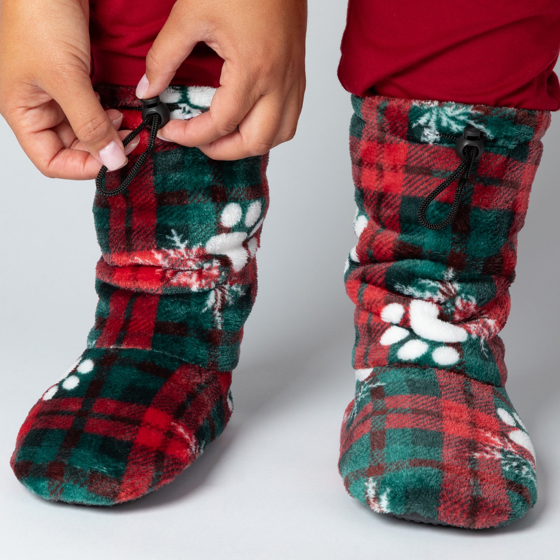 Red and green plaid slipper booties with paw prints and snowflakes, with adjustable toggles on top, against a white background.