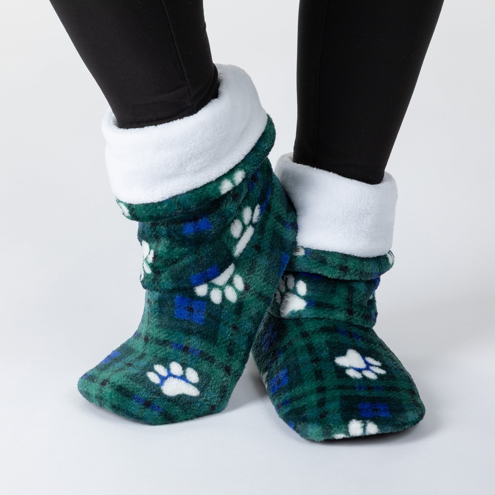 A pair of plush fleece slipper socks with a green and blue plaid pattern, white paw prints, and a white fleece cuff, shown on a person's feet wearing black leggings against a light gray background.