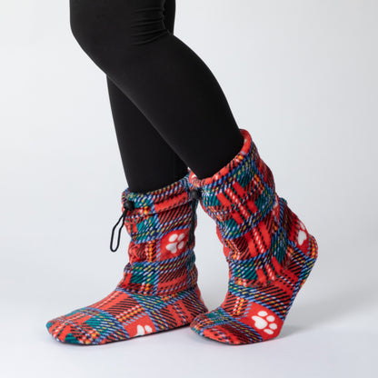 A person wearing red and multicolor plaid fleece booties with white paw print patterns, paired with black leggings, shown from the calves down against a light gray studio background.