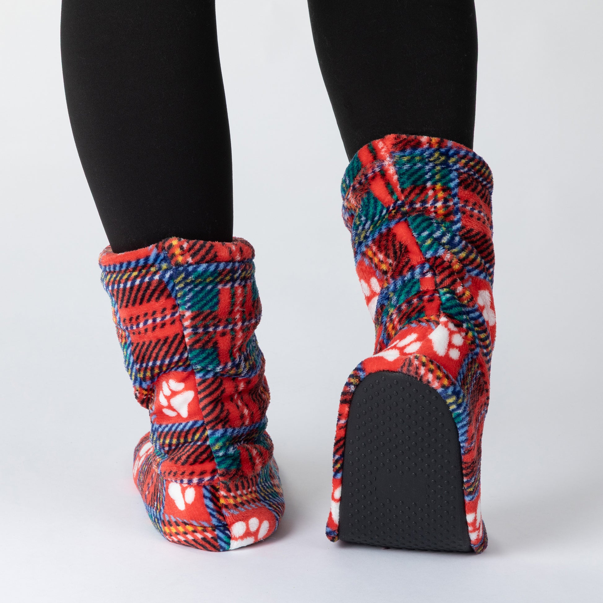 A pair of plush knit booties with a red, blue, and white plaid pattern and white paw prints, shown on a model wearing black tights against a light gray background.