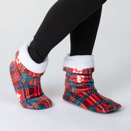 A pair of red and teal plaid slipper socks with white fleece trim, shown on a person's feet wearing black leggings against a light gray background.