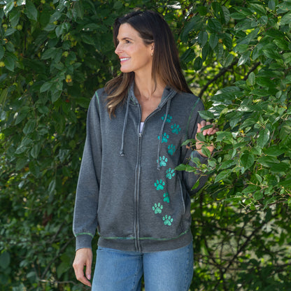 A woman wearing a dark gray zip-up hoodie with green paw print graphics on the front, standing outdoors in front of leafy green trees.