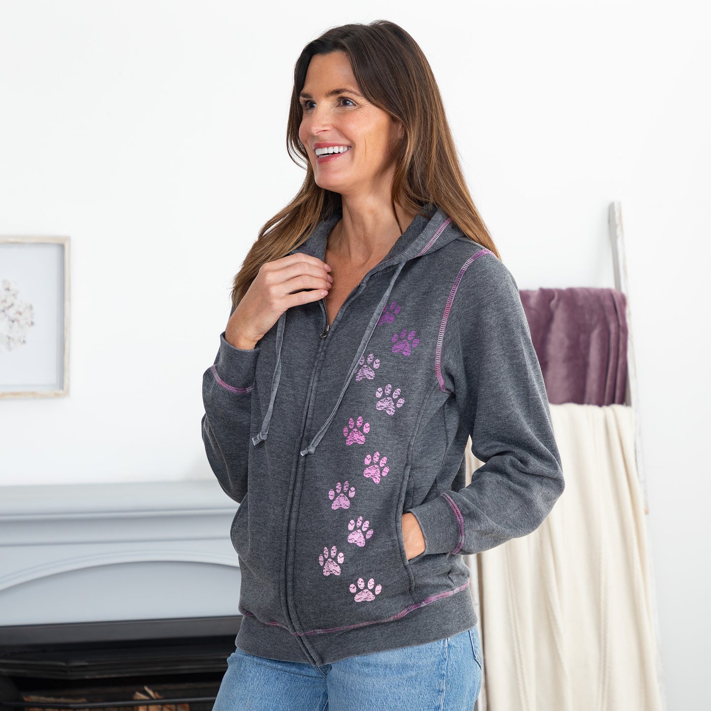 A woman wearing a dark gray zip-up hoodie with purple paw prints and purple trim, paired with blue jeans, standing in a brightly lit room.