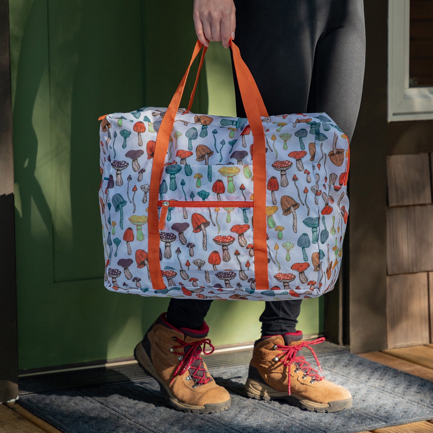 A person carrying a white duffel bag with various mushrooms designed on the fabric by the orange handle. The person is exiting a cabin in an outdoorsy setting. 