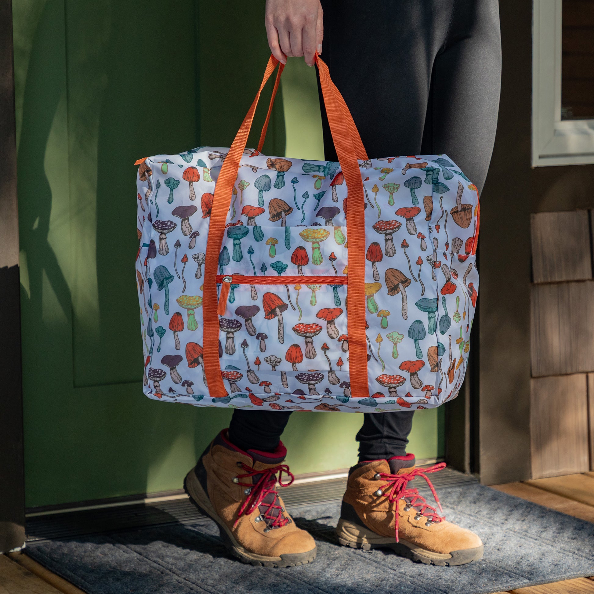 A person carrying a white duffel bag with various mushrooms designed on the fabric by the orange handle. The person is exiting a cabin in an outdoorsy setting. 