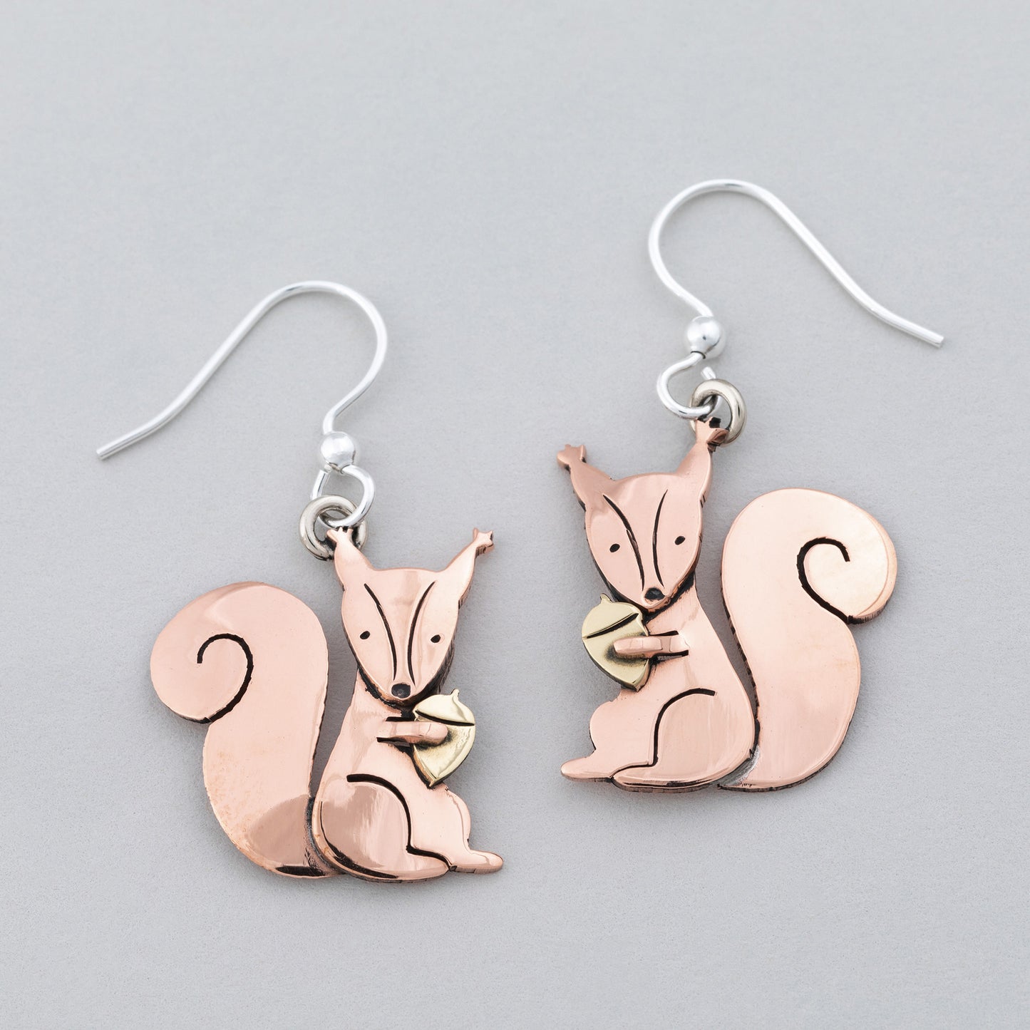 Copper squirrel holding a brass acorn on earrings on a gray background.