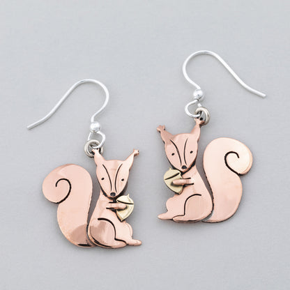 Copper squirrel holding a brass acorn on earrings on a gray background.