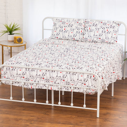 A white metal bed frame dressed with a white sheet and matching pillowcases featuring a repeating black cat print with small red bow-tie accents, set in a bright bedroom with wood flooring and a side table.