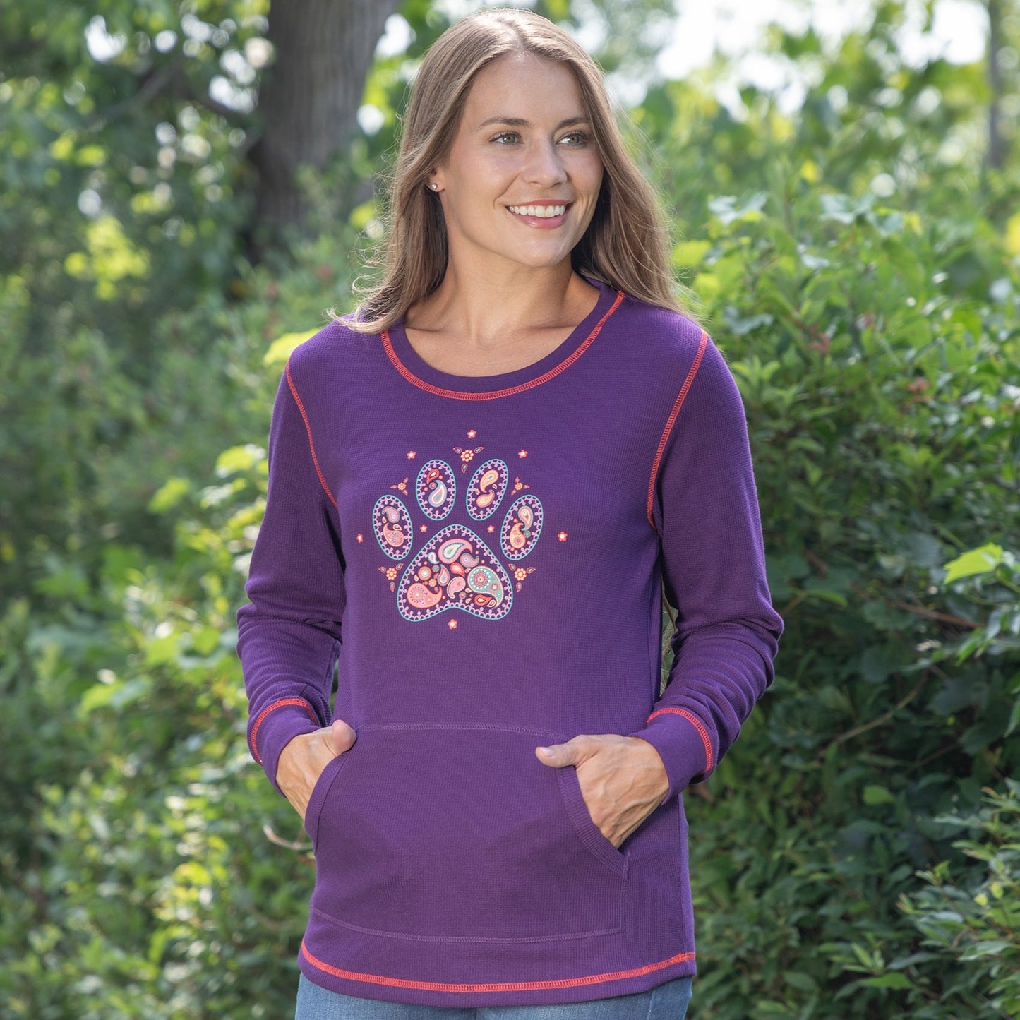 Woman wearing a purple thermal long sleeve top, with a paisley paw print design on the front, outdoors with trees in the background.