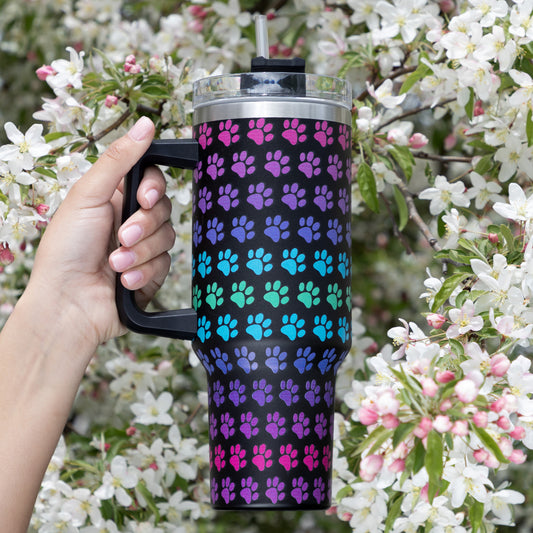 Hand holding a rainbow paw print patterned 40 oz drink tumbler, outside with flowers in the background.