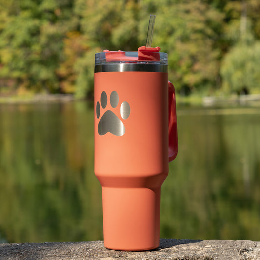 Coral 40 oz tumbler with a paw print design on a stone ledge by a body of water.