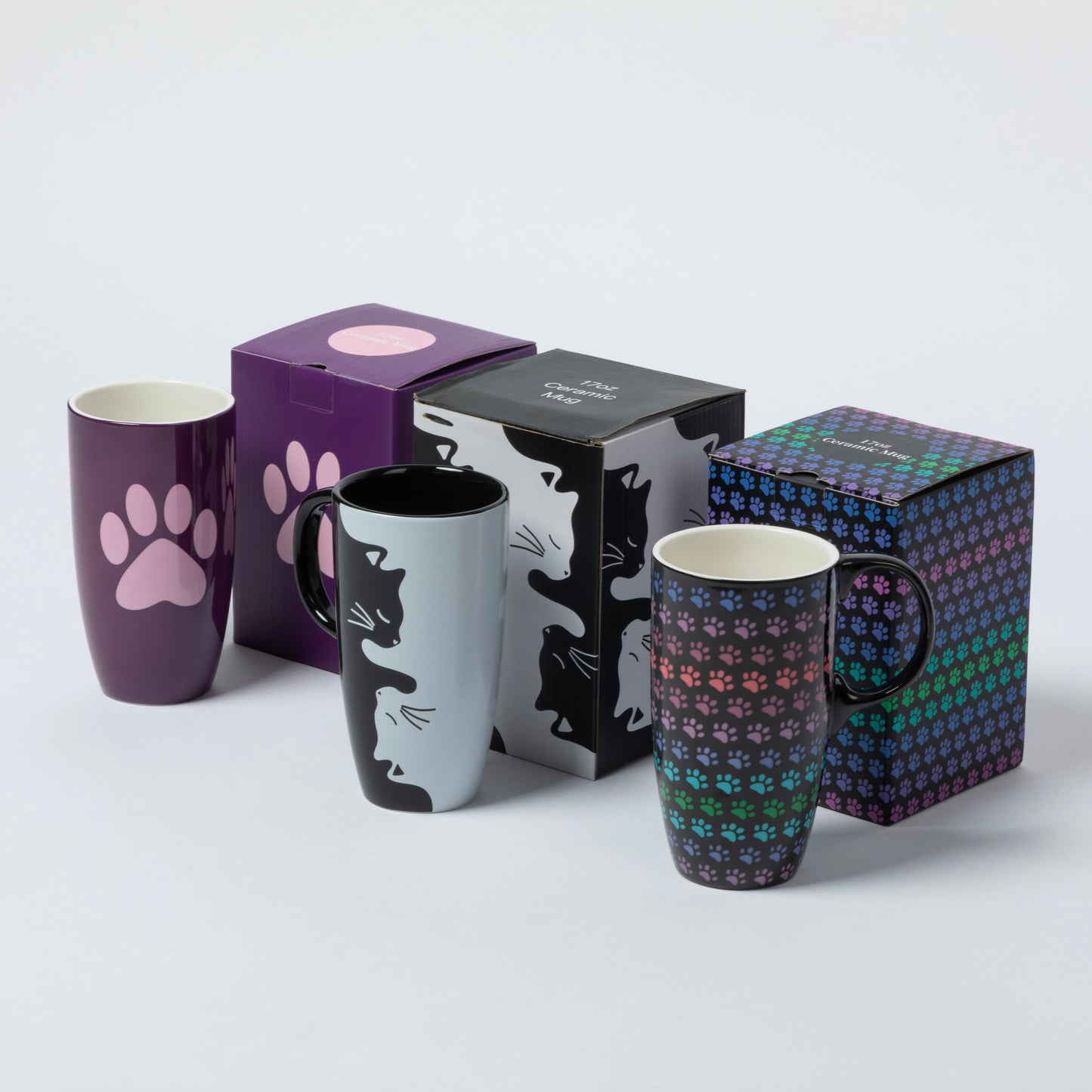 Three ceramic mugs with paw-themed designs are displayed alongside their packaging boxes on a light gray background. The mugs feature a purple paw print, a black-and-white cat silhouette, and a multicolor pattern of tiny cat paws.