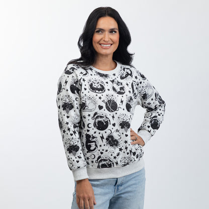 A woman wearing a light gray crewneck sweatshirt with a black celestial pattern of crescent moons, and stars, paired with light blue jeans, photographed against a light gray studio background.