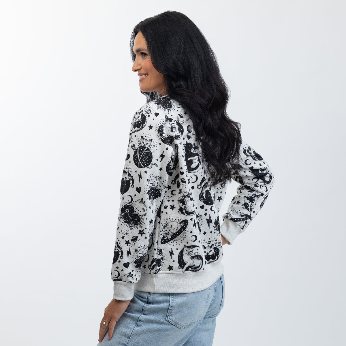 A woman wearing a light gray crewneck sweatshirt with a black celestial pattern of crescent moons, and stars, paired with light blue jeans, photographed against a light gray studio background.