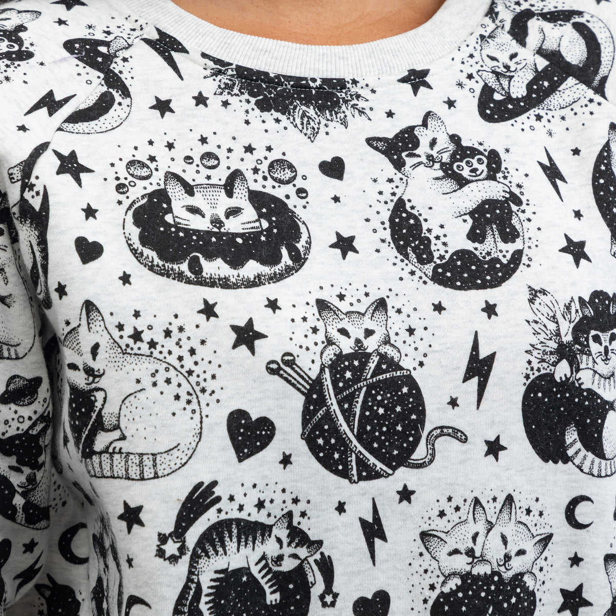 A close-up of a light gray crewneck sweatshirt featuring a black hand-drawn style print of cats, stars, hearts, and other celestial doodles.