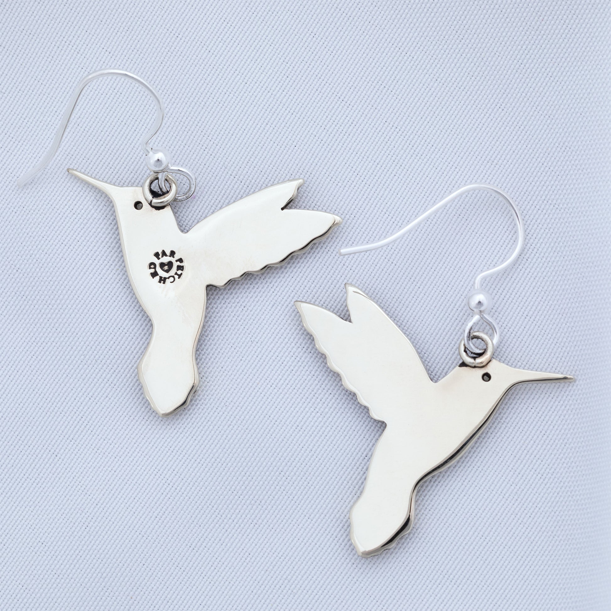 The backs of silver tone hummingbird earrings on a textured white fabric background.