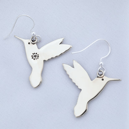 The backs of silver tone hummingbird earrings on a textured white fabric background.