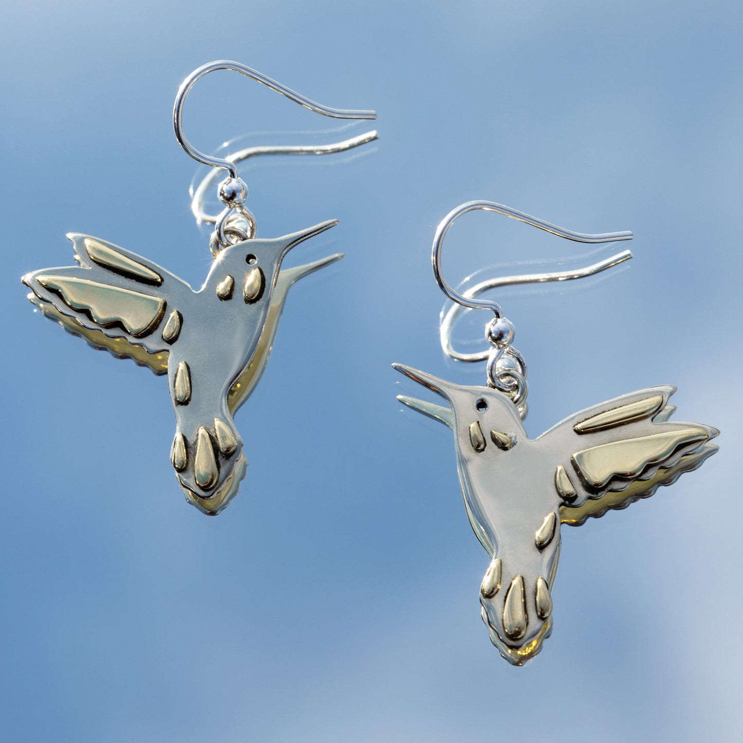 Silver hummingbird earrings with brass accents against a mirror reflecting the clear blue sky.