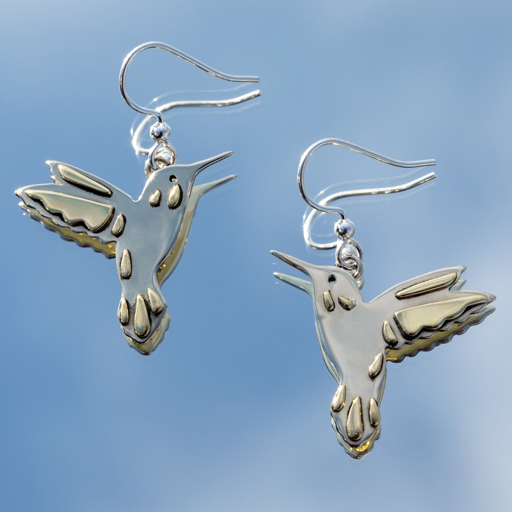 Silver hummingbird earrings with brass accents against a mirror reflecting the clear blue sky.