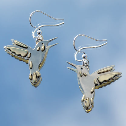 Silver hummingbird earrings with brass accents against a mirror reflecting the clear blue sky.