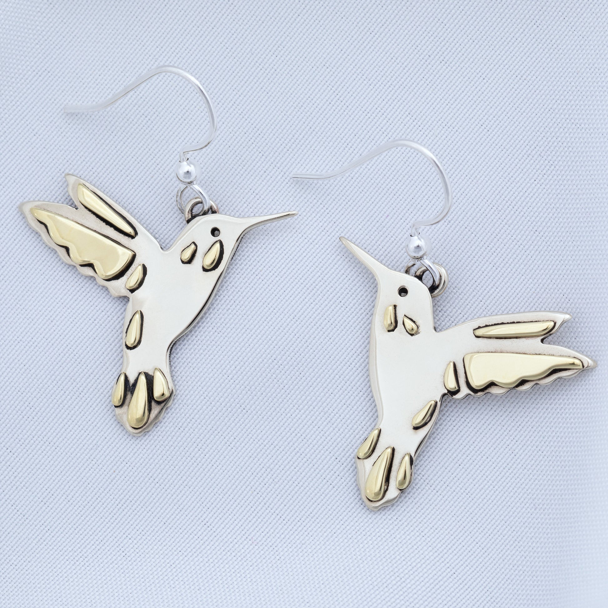 Silver hummingbird earrings with brass accents on a textured white fabric background.