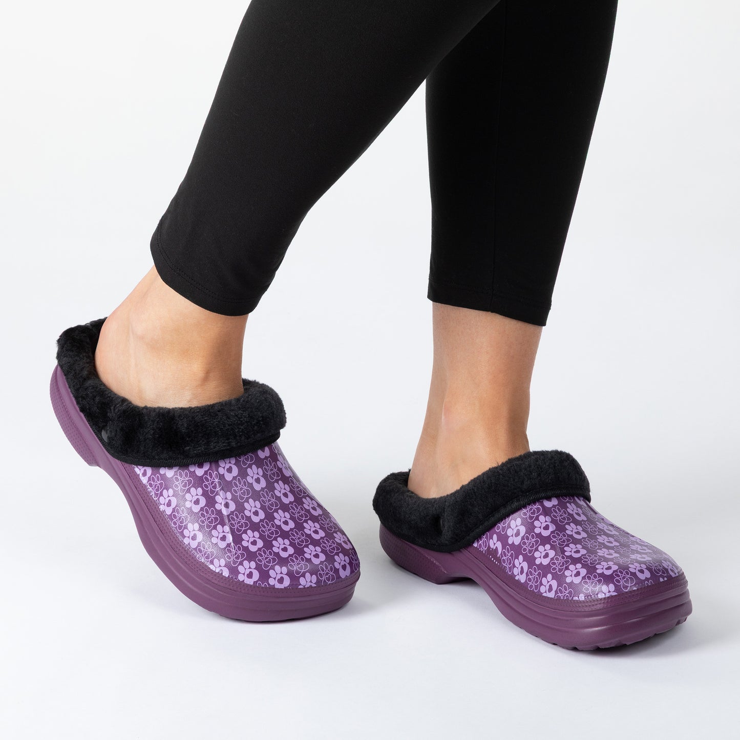 A person wearing slip-on clog-style slippers with a purple paw pattern, black faux-fur trim, and purple soles, shown against a light gray studio background.