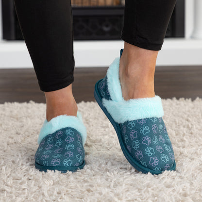 A person wearing slip-on slippers with a teal blue paw-print pattern and fluffy light blue trim, standing on a white shag rug.