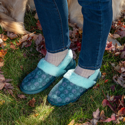 A person wearing blue slip-on slippers with a light blue fuzzy trim and a repeating paw print pattern, standing on grass outdoors with fallen leaves. The person is also wearing blue jeans and light-colored socks.