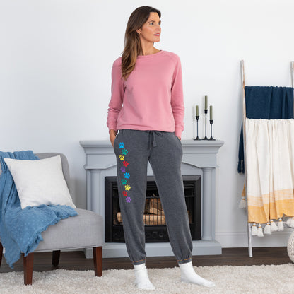A woman stands in a living room wearing a pink long-sleeve top and dark gray jogger sweatpants with colorful paw-print embroidery down one leg. The room has a fireplace, a chair with a blue throw, and a towel rack.