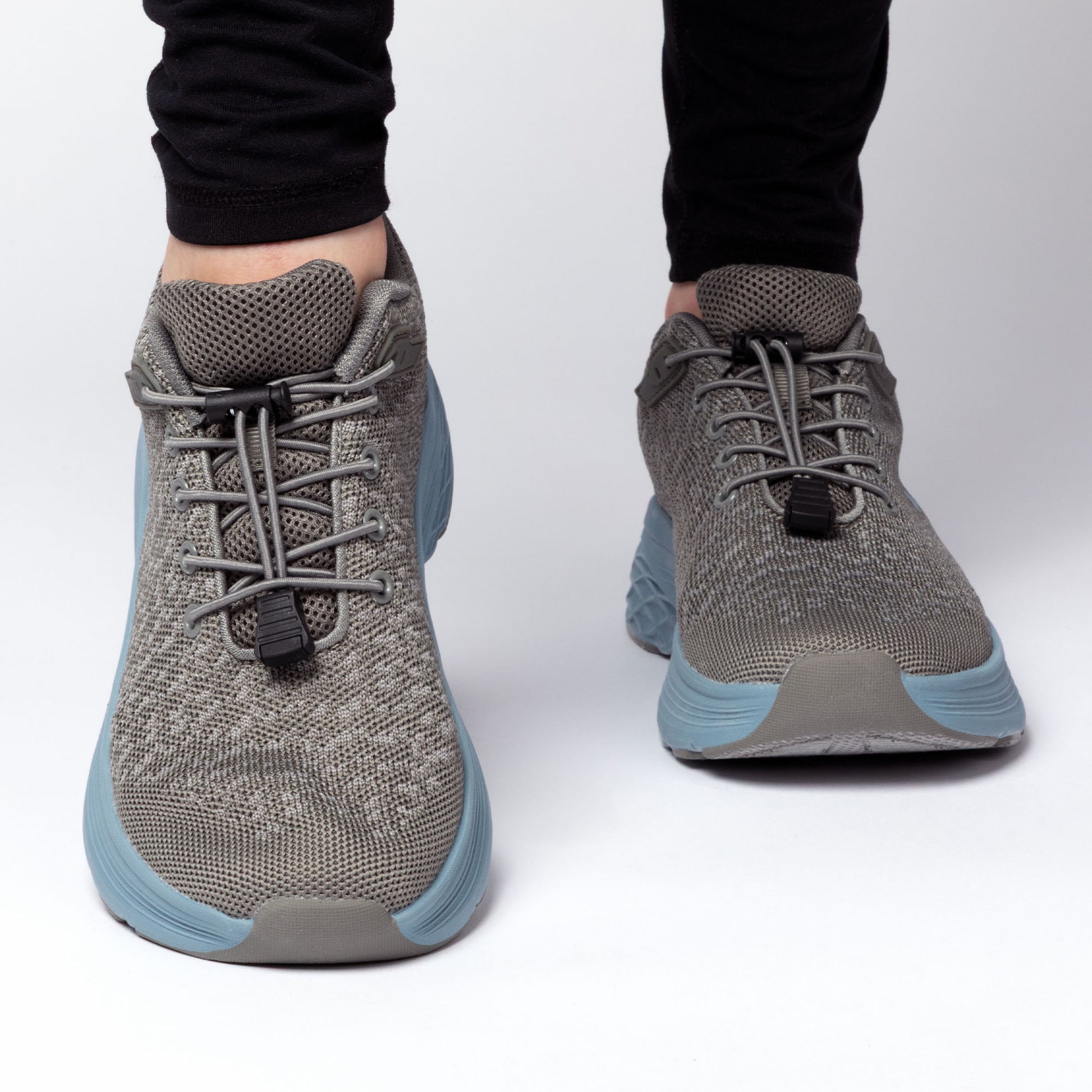 Gray sneakers with blue soles and gray bungee laces, worn by a person on a white background