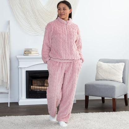 A woman wearing a cozy pink cable-knit fleece pajama set with a matching zip-up long-sleeve top and relaxed pants, standing in a bright living room with a fireplace, chair, and rug.